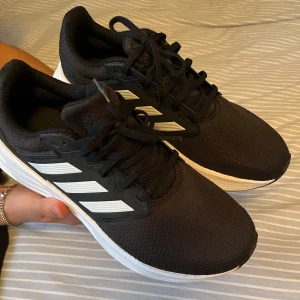 Svarta idrottsskor från Adidas - Säljer ett par svarta Adidas sneakers med vita ränder och snörning. Skorna har en bekväm sula och är perfekta för träning eller vardagsbruk. Tillverkade i syntetmaterial. Inte använda någonting bara testade pga av fel storlek. 