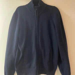 Mörkblå stickad kofta med dragkedja - Säljer en stilren mörkblå stickad kofta med dragkedja och ribbad krage. Perfekt för kyliga dagar och enkel att matcha med olika outfits.