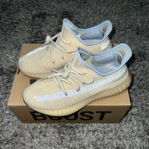 Yeezy Boost 350  - Säljer ett par beige Adidas Yeezy Boost 350 V2 med en stickad ovandel och snörning. Skorna har en genomskinlig sula och en glittrig detalj längs sidan. Perfekta för en stilren look.
