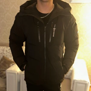 Svart dunjacka från Sail Racing - Snygg svart dunjacka från Sail Racing med huva och dragkedja. Jackan har en logotyp på bröstet och en på ärmen. Perfekt för kalla dagar. Vid snabb köp priset går o diskutera för att jag har redan en annan jacka 