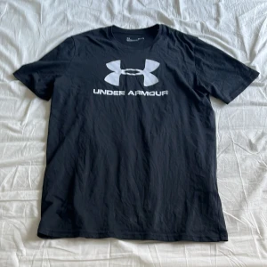 Svart t-shirt från Under Armour - Snygg svart t-shirt från Under Armour med deras ikoniska logga i vitt på bröstet. Perfekt för träning eller en avslappnad stil. Kortärmad och tillverkad i mjukt material för bästa komfort.