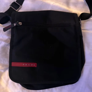 Svart axelväska från Prada - Snygg svart axelväska från Prada med justerbar rem och dragkedja. Väskan har en stilren design med en röd Prada-logga på framsidan och flera fack inuti för praktisk förvaring. Sparsamt använd.