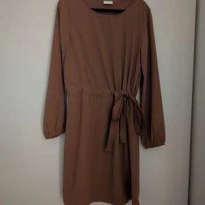 Brun klänning från Vila - Säljer en stilren brun klänning från Vila med smickrande knytband i midjan. Klänningen är perfekt för en elegant look. Oanvänd! ✨