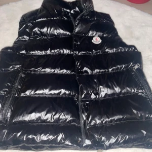 Svart dunväst från Moncler - Snygg svart dunväst från Moncler med glansig yta och dragkedja framtill. Västen har en hög krage och Moncler-logotypen på bröstet. Perfekt för kyligare dagar. Och priset kan diskuteras 