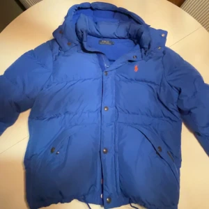 Blå dunjacka från Polo Ralph Lauren - Snygg blå dunjacka från Polo Ralph Lauren med huva och knappar. Jackan har en klassisk design med det ikoniska logotypen broderad i rött på bröstet. Perfekt för kyliga dagar. Jackan är i nyskick 