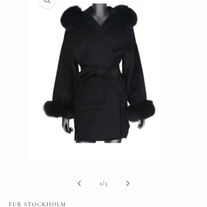 Svart kappa med pälsdetaljer - Elegant svart kappa från Fur Stockholm med lyxiga pälsdetaljer vid krage och ärmslut. Kappan har en omlott-design och knyts med ett bälte i midjan för en stilren look.