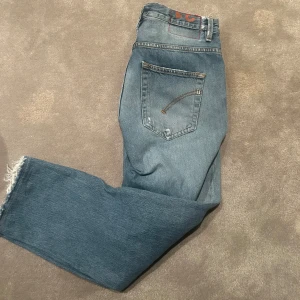 Dondup jeans med slitningar - Säljer ett par klassiska blå jeans med slitningar på knäna och fransiga benslut. De har en normal passform och är perfekta för en avslappnad stil. Jeansen har fem fickor och knappar i midjan.