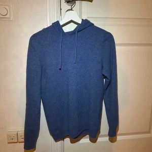 Blå merinoull hoodie - Säljer en mysig blå hoodie med långa ärmar och huva. Perfekt för kyliga dagar och avslappnade stunder. Den har en enkel design utan mönster eller detaljer.