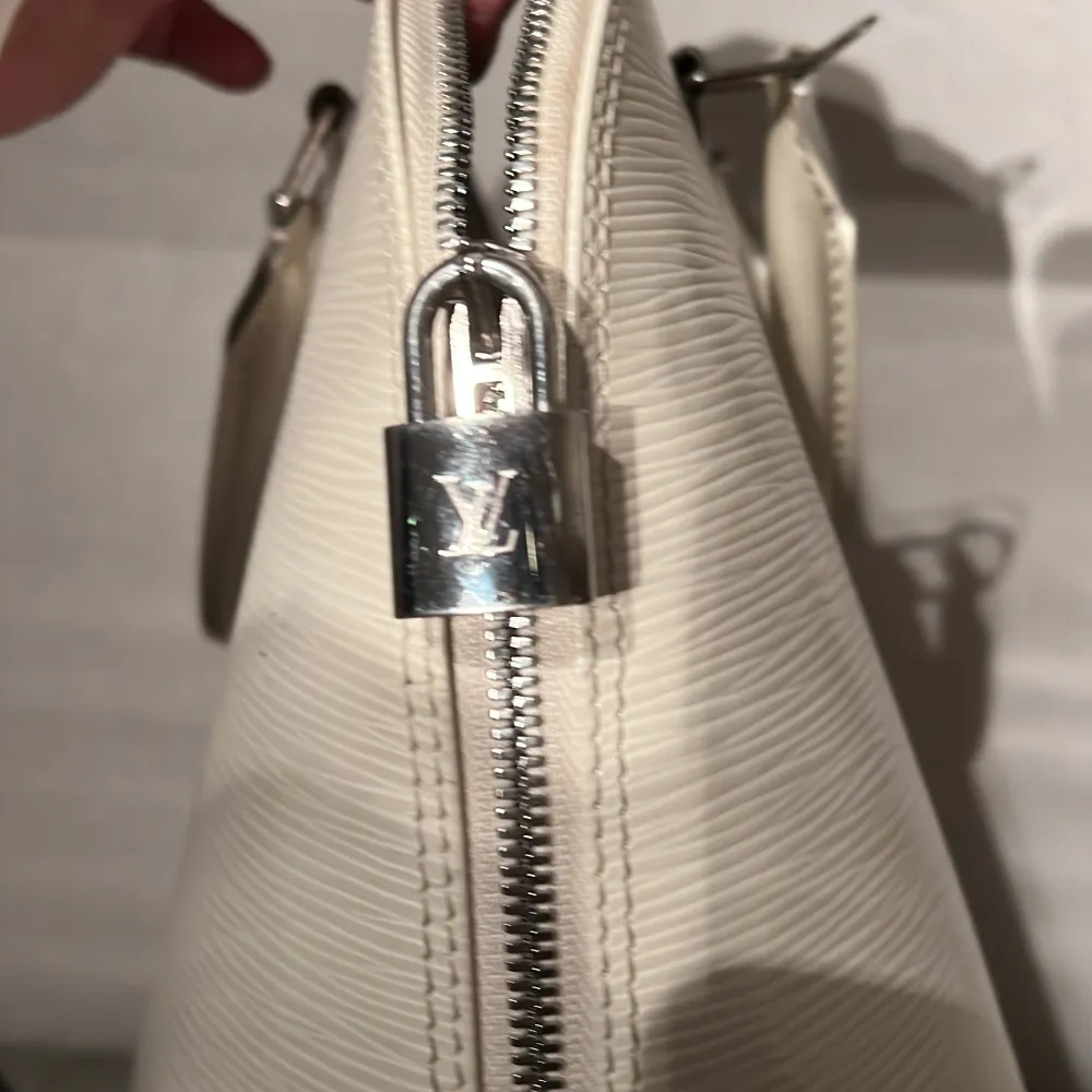 Säljer en elegant benvit klassisk ”Alma” från Louis Vuitton i Epi-skinn. Väskan har en stilren design med dragkedja och ett LV-lås . Väskor.