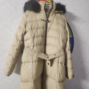 Tommy Hilfiger beige winter jacket, size L - En snygg beige vinterjacka med svart pälskrage. Jackan har en dragkedja och knappar framtill och en bälte i midjan. Jackan är fodrad och har två fickor framtill. Användt några gånger  Storlek L men passar M också 