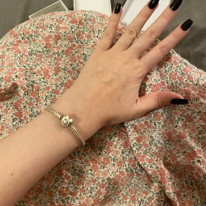 Silverfärgat armband med hjärtformad detalj från Pandora  - Säljer ett stilrent silver armband med en söt hjärtformad detalj från Pandora. Perfekt för både vardag och festliga tillfällen. Armbandet är enkelt att matcha med andra smycken och ger en elegant touch till din outfit. Man kan köpa till berlocker och jag säljer även några. Nypris 799kr, Storlek 18