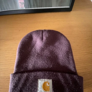 Lila mössa från Carhartt - Säljer en snygg lila mössa från Carhartt i nytt skick. Den är stickad och har en broderad logga framtill. Perfekt för att hålla sig varm under höst och vinter. One size fits all, så den passar de flesta. Grym accessoar för både vardag och fest!