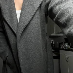 Grå kappa - Säljer en stilren grå kappa i mycket bra skick kappan är i  knä höjd. Den har en klassisk design med långa ärmar och en elegant krage. Perfekt för höst och vinter, och passar både till vardags och mer uppklädda tillfällen. Kappan är i ett mjukt material som känns bekvämt att bära.