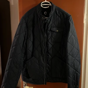 Mörkblå quiltad jacka - Säljer en mörkblå quiltad jacka i storlek L från Cubus. Jackan har en dragkedja framtill och en knappdetalj vid kragen. Den har också en bröstficka med knapp och två sidofickor. Perfekt för höst och vår!