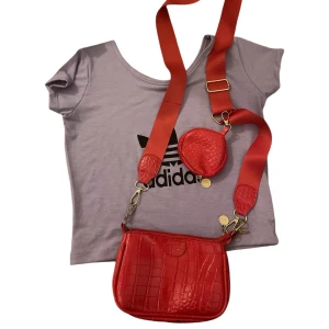 Röd axelväska med krokodilmönster - Snygg röd axelväska med krokodilmönster och matchande axelrem. Väskan har en liten rund väska fäst vid remmen, perfekt för småsaker. Den är i mycket bra skick och passar perfekt för både vardag och fest. En riktig färgklick som lyfter vilken outfit som helst!