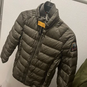 Olivgrön dunjacka från Parajumpers - Säljer en olivgrön dunjacka från Parajumpers med dragkedja och hög krage. Jackan har en quiltad design och en patch på ärmen. Perfekt för kyliga dagar.