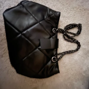 Svart quiltad axelväska med kedjehandtag - Snygg svart quiltad axelväska med kedjehandtag och röd insida. Perfekt för att ge en stilren touch till din outfit. Väskan har en elegant design med metallringar för handtagen.