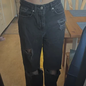 Svarta jeans med slitningar från NA-KD - Snygga svarta jeans från NA-KD med slitningar på knäna och en hög midja. De har en loose passform och klassiska femficksdetaljer. Perfekta för en avslappnad stil. Bekväm, och bra skick förutom att slitningarna har gått upp.