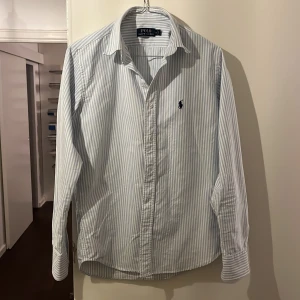 Randig skjorta från Polo Ralph Lauren - Snygg och klassisk ljusblå och vit randig skjorta från Polo Ralph Lauren. Skjortan har långa ärmar och en broderad logotyp på bröstet. Perfekt för en stilren look. 