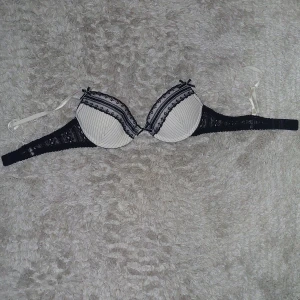 Svart och vit spets-bh - Elegant bh i svart och vit med spetsdetaljer och små rosetter. Den har justerbara axelband och en klassisk knäppning bak. Perfekt för en stilren look.