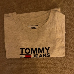 Grå t-shirt från Tommy Jeans - Säljer en stilren grå t-shirt från Tommy Jeans med deras ikoniska logga tryckt på bröstet. Perfekt för en avslappnad look.