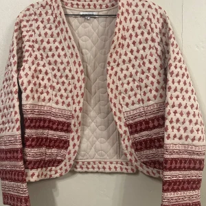 Beige och röd quiltad jacka från Glamorous - Säljer en unik quiltad jacka från Glamorous i beige med rött mönster. Jackan har långa ärmar och ett detaljerat mönster som ger en bohemisk känsla. Perfekt för att ge din outfit en färgklick och stil.