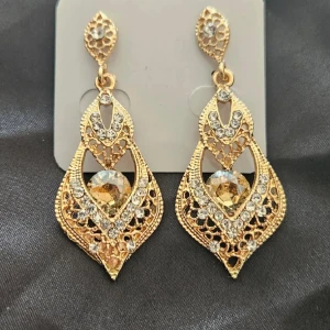 Guldglittriga örhängen med stenar - Eleganta guldglittriga örhängen med intrikata detaljer och gnistrande stenar. Perfekta för att ge en lyxig touch till din outfit. De har en droppformad design med en stor central sten omgiven av mindre stenar.