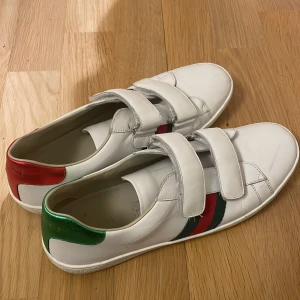 Vita sneakers med kardborreband från Gucci - Snygga vita sneakers från Gucci med kardborreband och klassiska ränder i grönt och rött på sidan. Skorna har en röd och en grön detalj på hälen med Gucci-logga. Perfekta för en stilren look. Kartong finns