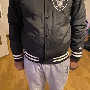 Svart bomberjacka med NFL-logga - Snygg svart bomberjacka från New Era med broderad NFL-logga och Raiders-emblem på bröstet. Jackan har vita ribbade muddar med svarta ränder och knappar framtill. Perfekt för sportfans