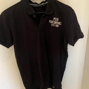 Svart pikétröja från Peak Performance - Snygg svart pikétröja från Peak Performance med vit text på bröstet. Tröjan har en klassisk krage och knappar framtill. Perfekt för en stilren look.