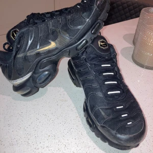Svarta TN Nike Air sneakers - Säljer ett par svarta TN Nike Air sneakers med snörning och en unik sula i svart och guld. Perfekta för en sportig look.