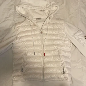Vit dunjacka med huva från Moncler - Snygg vit dunjacka från Moncler med huva och dragkedja. Jackan har långa ärmar och praktiska fickor med dragkedjor. Perfekt för kyligare dagar.