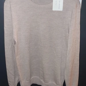 Beige merinoulltröja från Tiger of Sweden - Säljer en stilren beige tröja i mjuk merinoull från Tiger of Sweden. Perfekt för kyligare dagar med sin långärmade design och klassiska passform. En tidlös baströja för garderoben. Sprillans ny så inga defekter