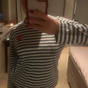 Randig tröja från Comme des Garçons - Säljer en randig tröja från Comme des Garçons med blå och vita ränder. Tröjan har ett rött hjärta med ögon på bröstet och långa ärmar. Perfekt för en stilren look!