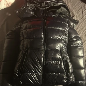 Svart pufferjacka med huva - Snygg svart pufferjacka med glansig yta och praktisk huva. Jackan har dragkedja framtill och två fickor med dragkedjor. Perfekt för kyliga dagar.