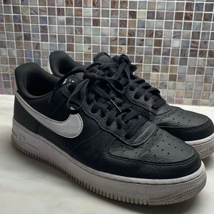 Svarta Nike Air Force sneakers - Säljer ett par klassiska svarta Nike Air Force sneakers med vit sula och vit swoosh. Skorna har snörning och är tillverkade i skinn. Perfekta för en stilren look.
