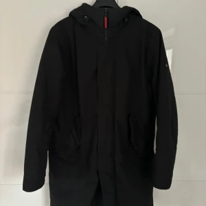 Svart vinterjacka från Elvine - Snygg svart parkajacka från Elvine med Thermore-foder för extra värme. Jackan har en stilren design med dragkedja och huva, perfekt för kyliga dagar. Insidan har ett diskret camouflage-mönster.