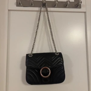 Svart quiltad axelväska med kedja - Snygg svart axelväska i quiltat mönster med en stilren kedja som axelrem. Väskan har en stor dekorativ ringdetalj på framsidan som ger den en modern touch. Perfekt för att lyfta vilken outfit som helst!