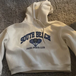 Vit hoodie från 157 - Säljer en vit hoodie från 157 med trycket 'South Beach Padel Sports Club' i blått. Hoodien har en klassisk design med dragsko i huvan och ribbade muddar vid ärmslut och nederkant. Perfekt för en avslappnad stil.