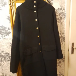 Svart kappa från Busnel - Elegant svart kappa från Busnel med guldknappar och hög krage. Kappan har långa ärmar och två stora fickor framtill. Perfekt för en stilren look. Ordinarie pris ca 8000