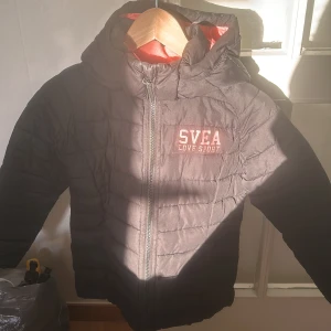 Svart pufferjacka från Svea - Säljer en svart pufferjacka från Svea med texten 'Love Sport' på bröstet. Jackan har en huva och dragkedja framtill. Insidan är fodrad i en kontrasterande rosa färg för extra värme och stil. Obs har ett litet hål på baksida arm, du ser det på sista bilden. ”Går att diskutera pris”