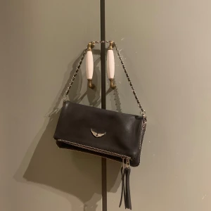 Svart axelväska från Zadig & Voltaire - Snygg svart axelväska från Zadig & Voltaire med kedjeaxelrem och dekorativt emblem. Väskan har en dragkedja och är perfekt för att bära dina nödvändigheter med stil.