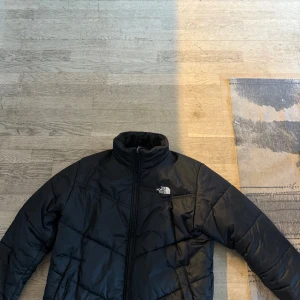 Svart dunjacka från The North Face - Säljer en svart dunjacka från The North Face. Jackan har en hög krage och dragkedja framtill. Perfekt för kyliga dagar med sin quiltade design som håller värmen. En klassisk och stilren jacka för alla tillfällen.