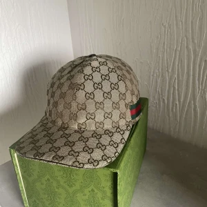Beige keps från Gucci - Snygg beige keps från Gucci med det klassiska GG-mönstret. Kepsen har en justerbar rem baktill och en röd-grön randdetalj på sidan. Perfekt för att ge din outfit en lyxig touch.