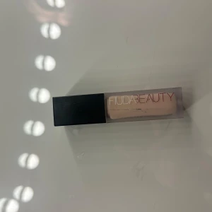 Concealer från Huda Beauty - Säljer en concealer från Huda Beauty med en ljus nyans. Den kommer i en rektangulär förpackning med svart lock och transparent kropp som visar färgen. Perfekt för att täcka ojämnheter och ge en jämn hudton.