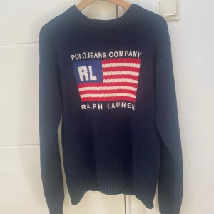 Mörkblå stickad tröja från Ralph Lauren - Säljer en snygg mörkblå stickad tröja från Ralph Lauren med ett stort amerikanskt flaggmotiv och texten 'Polo Jeans Company' på framsidan. Tröjan har långa ärmar och är tillverkad i 100% bomull. Perfekt för en stilren och avslappnad look. Äldre modell.