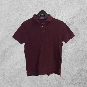 Vinröd pikétröja från Ralph Lauren - Snygg vinröd pikétröja från Ralph Lauren med klassisk krage och knappslå. Den har en liten broderad logotyp i blått på bröstet. Perfekt för en stilren och avslappnad look.