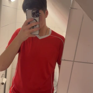 Röd fotbollströja med vita detaljer - Säljer en röd fotbollströja med vita detaljer och V-ringning. Tröjan är kortärmad och har en sportig design med vita linjer längs sidorna och ärmarna. Perfekt för träning! 