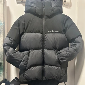 Svart dunjacka från Sail Racing - Snygg och varm svart dunjacka från Sail Racing med dragkedja och hög krage. Perfekt för kyliga dagar med sin stilrena design och funktionella detaljer.