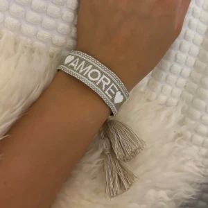 Beige armband med texten 'AMORE' - Charmigt beige armband med texten 'AMORE' och hjärtdetaljer. Armbandet har dekorativa tofsar som ger en bohemisk känsla. Perfekt accessoar för en avslappnad stil.